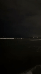 Nighttime waves on a dark coastline, distant lights on the horizon
