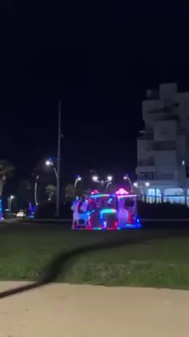 At night, the video pans across a public square or park. The ground is a mix of sand and grass. In the background, there are palm trees and a tall white building with stacked, modular designs. Several rides that look like carriages are arranged on the grass, each brightly lit with multi-colored LED lights. Some people are standing near the rides, possibly waiting their turn or supervising children. Street lights illuminate the area, and some cars are visible in the distance. The sky is dark.