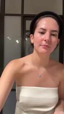 The video shows a woman with fair skin washing her face in a bathroom. She is wearing a white strapless top and a black headband, and she has a gold necklace and bracelet. She rubs her hands together with water and then brings them up to her face to wash it. Behind her is a dark-framed glass partition with multiple panes. The bathroom is well-lit.