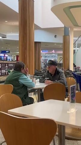 The video shows two elderly people, one male and one female, sitting at a table in what appears to be a cafe or food court. The male, on the right, wears a grey jacket and a black baseball cap. He is holding a cup and looking at the female. The female, on the left, wears a green vest over a blue shirt and is looking at the male. Both are seated at a white table with chairs around it. Behind them, the environment of the cafe/food court is visible, with other tables, chairs, pillars, and storefronts in the background. The lighting is natural and appears to be from overhead. There is a clear plastic display stand on the table in the foreground.