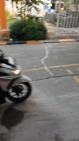 A motorcycle rider wearing a white helmet and a black jacket is seen from the side, riding a white motorcycle. The motorcycle has large black bags attached to the rear, and two large brown bags are strapped to the sides. The rider is wearing blue jeans. The motorcycle is moving from left to right, passing through a gate. The gate appears to be an automated barrier gate, with a white arm that is in the process of rising. Red and white flags are visible in the background, along with trees and other foliage. The ground is paved and appears to be wet in places. A security booth with a sign that says "SECURITY" is visible on the right side of the frame. Another person, partially visible in the background, is standing near the gate. The video is a bit blurry due to motion.