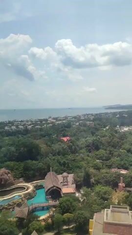 The video presents a high-angle view of a resort or theme park on a bright, partly cloudy day. A body of water, likely the ocean, is visible in the background, stretching along the horizon. The foreground is dominated by a dense, green canopy of trees, interspersed with buildings of various designs. A prominent castle-like structure with multiple spires and turrets can be seen amidst the greenery. In the lower-left corner, a swimming pool complex with artificial rock formations and a thatched-roof building is visible. The camera slowly pans right, revealing more of the complex and the surrounding landscape.