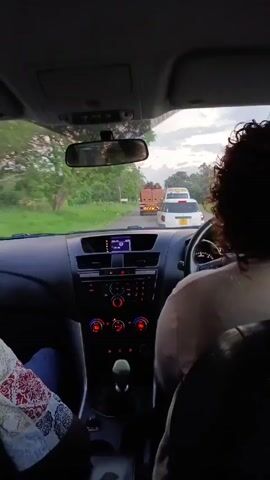 Here is a description of the video:
The video is shot from inside a car driving on a paved road, looking forward through the windshield. A person with curly hair is driving, and another person is sitting in the passenger seat. A truck and a white SUV are visible ahead on the road. Trees line both sides of the road, and the sky is overcast. The car's dashboard is visible, with the radio, air conditioning controls, and gear shift in clear view.
