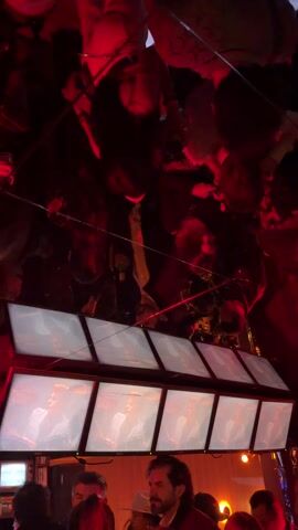The scene takes place in a bar or event space with a lively atmosphere. A bank of seven screens displays a person singing or performing. The screens are mounted low, at head height of the people in the foreground, and angled slightly upward. The foreground is filled with people, some of whom are looking at the screens and others who are obscured. The space is lit with red and orange lighting, creating a warm and energetic atmosphere. Above the screens, a mirrored ceiling reflects the crowd, creating a sense of a packed and energetic space. There are hanging lights or decorations visible in the reflection.