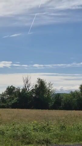The video shows a rural landscape under a bright blue sky with scattered clouds. In the foreground, there is a field of dry grass and some bushes, with a line of trees forming a backdrop across the middle of the frame. In the sky, a contrail from an airplane is visible as a long white line against the blue. The camera pans slightly to the right, revealing a traffic sign warning of winding roads. The scene is calm and peaceful, with no people or other prominent objects. The overall impression is of a sunny day in the countryside.
