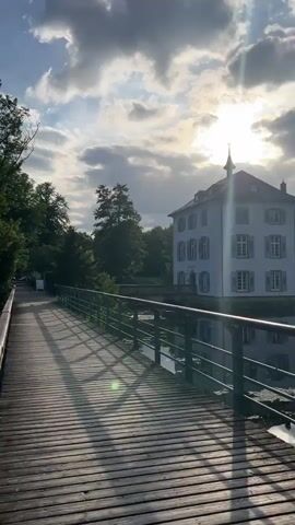 A wooden footbridge with a dark railing extends from the left foreground into the middle ground of the video. Sunlight casts long shadows from the railing across the bridge's wooden planks. To the right of the bridge, a calm body of water reflects the sky and surrounding trees. A white building with a pale facade and shutters is visible on the far bank of the water, partially obscured by trees. The sky above is blue with scattered white clouds, and the sun is partially visible, creating a bright glare in the upper right corner. Trees with lush green foliage line the left side of the footbridge and extend behind the building on the right. In the water, a white swan is visible swimming near the bank on the left, and a group of ducks are visible on the water in the middle ground. The scene depicts a tranquil waterside environment with a bridge leading towards a building.