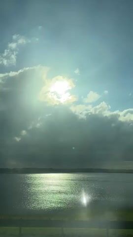 The video shows a scenic view of a lake from the perspective of a moving vehicle. The sun shines brightly through a mix of white and grey clouds in a blue sky, creating sunbeams over the water. The water's surface reflects the sunlight, causing a sparkling effect. The opposite shore is visible in the distance. A roadside barrier is visible, adding to the vehicle perspective. The video seems to have been recorded during daylight.