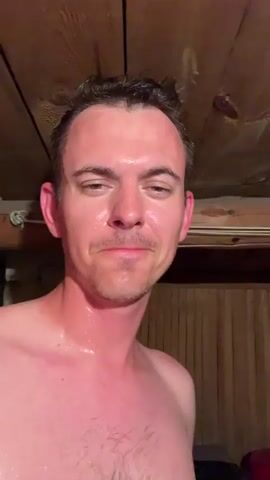 A man with short, dark hair and a mustache is looking into the camera. He is shirtless and appears to be sweating. He has reddish skin, possibly from heat or sunburn. The background shows a wooden-paneled wall and a wooden ceiling with exposed beams. There is also a red bucket visible to the right of the man. The man makes facial expressions, including a slight smile and an open-mouthed gesture. He briefly touches his chest.