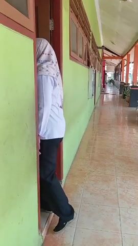 A person wearing a white uniform and a hijab walks through a doorway on the left side of the frame. The person is moving from inside a room to a hallway. The hallway has a tiled floor and a light green wall. There are windows along the wall. The doorway has a red frame and a brown door. The person's lower body, including black pants and shoes, is visible as they exit the room. The person is partially obscured by the door.