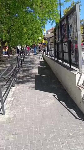 The video shows a view from a tram station platform. The platform has a paved surface and is lined with metal railings. Steps lead down from the platform to the street level. Across the tracks, there is a red brick structure with arches. The tram station sign reads "Burjassot-Godella". Trees line the area surrounding the station. Several people are walking on the street near the station entrance, while others can be seen standing or sitting on the platform itself. The sky is clear and blue. Overhead tram wires are visible.