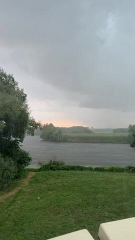 A video captures a natural landscape with a river, trees, and grass under a cloudy sky. Rain is falling, creating ripples on the water's surface. The overall atmosphere is overcast and somber.