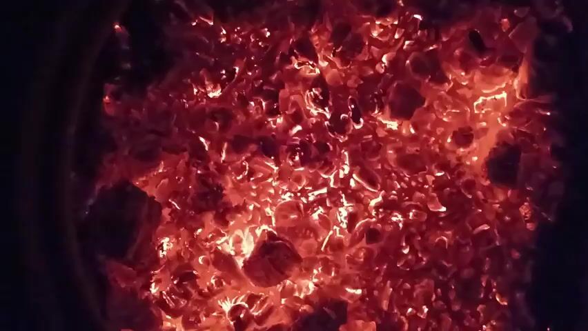 A close-up, overhead view of glowing red-hot coals in darkness. The coals are irregular in shape and size, piled together. The light emitted from the coals illuminates the immediate surroundings with a warm, orange-red glow.
