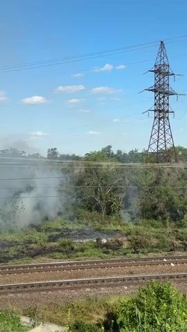 The video captures a daytime scene with a bright blue sky and scattered white clouds. In the foreground, there are railroad tracks with ballast. Beyond the tracks, a grassy area is visible, with a large plume of white smoke rising from it, indicating a fire. To the right of the smoke, a tall, lattice-style electrical tower stands against the sky. The surrounding vegetation appears to be a mix of green bushes and trees. The camera is positioned from a slight elevation, looking across the tracks towards the smoky area.