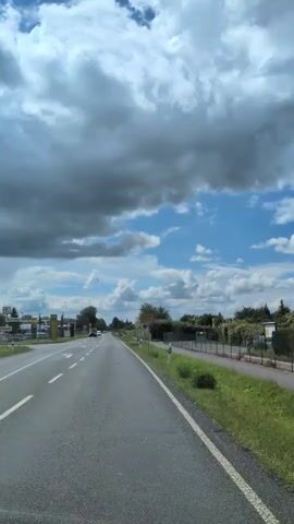 A point-of-view shot captures a view from a moving vehicle on a two-lane asphalt road. The road is bordered by a paved shoulder and a grassy verge, with a hedgerow running along the right side. In the distance, several cars are visible, and there are buildings and trees on the left side of the road. The sky is partly cloudy, with dramatic dark clouds interspersed with patches of blue sky. A yellow sign with German text is visible on the right side of the road at the beginning of the clip.