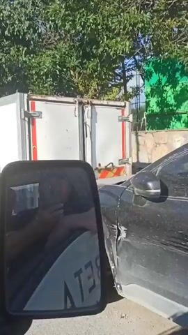 A white van with "OKUL TAŞITI" printed on the back drives down a street. The van has yellow and black diagonal stripes on the rear. There are trees and a blue car in the background.