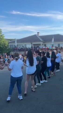 A group of women and girls are holding hands and dancing in a circle in the middle of a street. The women are wearing casual clothing such as t-shirts and jeans, while the girls are dressed in more youthful attire. Behind them, a building with a brown roof and white walls is visible, with people sitting at tables on a patio in front of it. The sky is blue with some clouds, and the sun is shining. The overall atmosphere appears to be festive and communal, possibly a celebration or a cultural event.