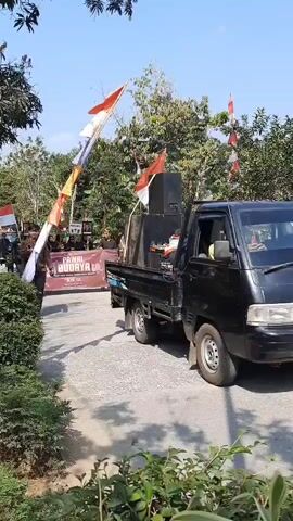 A black pickup truck with large speakers in the back is driving on a paved road. The truck is adorned with Indonesian flags and colorful banners. In front of the truck, a group of people is marching, also carrying flags and banners. One man in the foreground, wearing a patterned brown shirt and black pants, is walking and holding a microphone. The environment appears to be outdoors with trees and foliage visible. The sky is clear and blue.