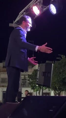 A man in a suit stands on a stage, illuminated by stage lights. He gestures with his hands as he speaks or sings. In the background, there are trees and buildings. A speaker is visible on the stage.
