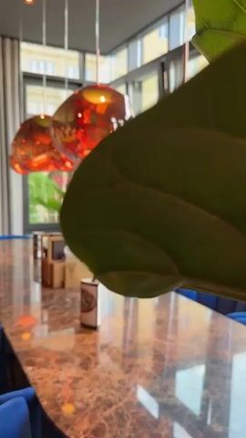The video shows a close-up, low-angle shot of a large, glossy, marble-patterned table in a dimly lit restaurant. The table is set with blue chairs. Several abstract, orange-hued glass pendant lights hang above the table, casting warm reflections on the table's surface. A green plant in a white pot sits on the table, along with a wooden organizer containing menus and napkins. The background, viewed through large windows, shows a blurred cityscape with buildings and trees. The camera slowly pans across the table, focusing on the lights and the table's reflective surface. The overall mood is sophisticated and modern.