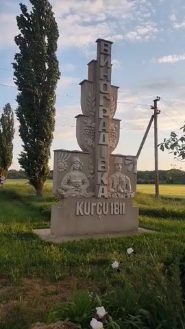 The video opens with a medium shot of a concrete monument marking the entrance to Vinogradivka, Ukraine. The monument has carved images of people and the year 1811. The camera pans right to reveal a small, open-air structure with a cross on top. The structure has a gray roof, white columns, and a brick base. Inside, there is a crucifix. White stairs lead up to the structure. The scene is bathed in the warm light of the setting sun. The area around the structure is decorated with flowers. The sky is blue with scattered clouds. A power line can be seen in the background.