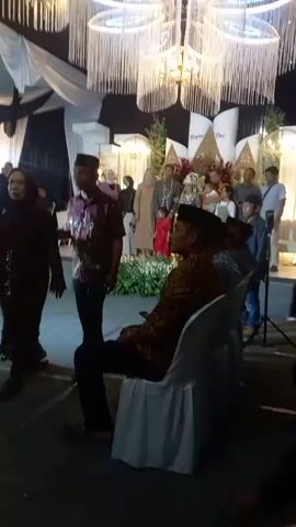 A wedding reception is underway, with guests gathered around the wedding party on a raised platform. The bride and groom are in the center of the platform, surrounded by family and friends. The room is decorated with white drapes, chandeliers, and floral arrangements. Guests are seated in white-covered chairs, with some standing and conversing. One man is seated in the foreground, facing away from the camera. Another man is standing to the left of the platform, facing the wedding party. The lighting is dim, with spotlights illuminating the platform.