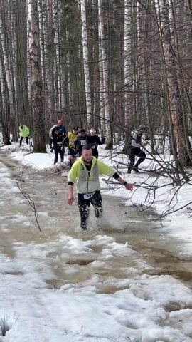 The video shows a group of people running in a snow-covered forest. In the foreground, a man wearing a bright yellow jacket and black leggings is running through a shallow stream of water. He is wearing a number "498". Other runners can be seen behind him, some closer to the stream and some on the snowy banks. The trees in the forest are mostly bare, and there are patches of snow on the ground. The sky is overcast.