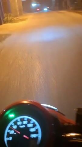 A first-person point-of-view video shot at night from a moving motorcycle. The camera is focused on the road ahead and the motorcycle's dashboard. The dashboard shows the speedometer, which indicates a speed of approximately 40 km/h. The road is paved and appears wet, with streetlights and the headlights of other vehicles visible in the distance. The rider's shadow is cast on the road. There are other motorcycles and people visible in the distance.