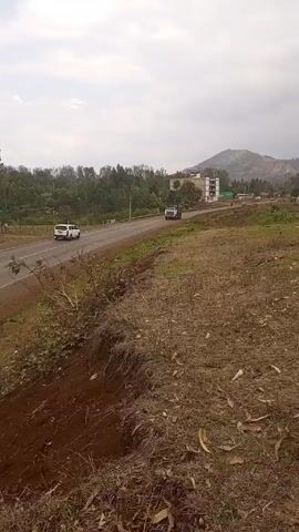 The video shows a white and dark gray dump truck moving on a road in a rural area. There's another vehicle driving in the opposite direction and a building visible in the background, as well as trees and rolling hills. The camera is static, and the focus is on the truck as it passes by. The area around the road is covered in dry vegetation.