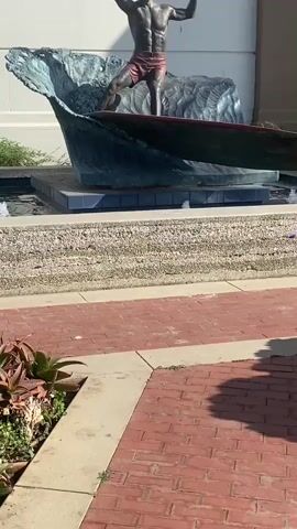 A bronze statue of a surfer on a wave is in the foreground. The statue is in a fountain with a gravel base and red brick paving. Behind the statue, a white building with a brown column and green plants is visible. The building has a marquee that reads "PLAYHOUSE" and lists upcoming shows. The sky is blue with clouds. The video pans from the statue to the building.
