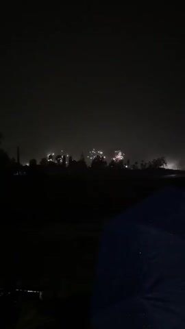 The video shows a nighttime scene with fireworks. The foreground is dark, with the silhouette of a blue object, possibly a tarp or umbrella, in the lower right corner. Beyond that, a dark landscape is visible, with trees silhouetted against the sky. In the distance, fireworks are exploding, illuminating the sky with bursts of light in various colors, including red and white. The fireworks appear to be set off over a wooded area or town. The sky is dark and hazy, suggesting a cloudy or misty night. The video captures the visual spectacle of a fireworks display.