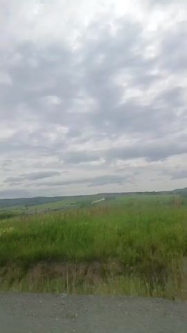A still video displays a cloudy sky over a green field and a dirt road. The field is covered in tall grass and some scattered structures are visible in the distance. The road is in the foreground.