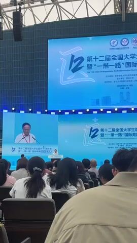 The video captures a large indoor event, likely a conference or competition, with a prominent stage and a large screen displaying text and graphics. A speaker, an adult male wearing a white collared shirt and a lanyard, stands at a podium on the left side of the stage, addressing the audience. The large screen shows the title of an event in both Chinese and English: "The 12th China Undergraduate Medical Innovation Tournament & The Belt and Road International Competition 2026 Finals." Below this, details about the organizers and dates (2026-08-14-16) are visible. The lower part of the screen also displays the event's title, mirrored. The audience is seated in rows, facing the stage, with the camera positioned behind some of the attendees, showing the backs of their heads and shoulders. The environment appears to be a modern convention center or auditorium with a high ceiling and ample lighting.