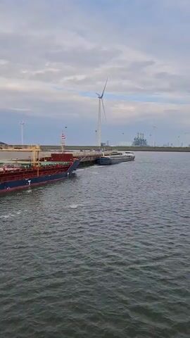 The video shows a large barge-type cargo ship named "Kelly C" in an industrial port setting.  The ship, primarily dark blue with a red hull, is docked at a quay lined with industrial structures including what appears to be a grain loading facility and shipping containers.  A white-painted loading arm extends towards the ship from the quay.  In the background, there's a wind turbine.  The water is choppy and dark. The viewpoint is from another vessel, with a small section of its black railing visible in the foreground.  The sky is overcast with gray clouds.