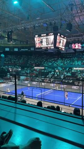 This is a video taken from an elevated spectator seat looking down at an indoor padel tennis match. A large, scoreboard is visible above the court, displaying player names and scores. The court itself is blue, enclosed by glass walls and a net dividing the two sides. Several players are on the court, actively playing the game. A large crowd fills the stands surrounding the court, illuminated by bright overhead lighting that casts a turquoise hue. In the foreground, the legs and feet of a person are visible, suggesting the video is recorded from a spectator's perspective. The atmosphere appears to be a lively sporting event.