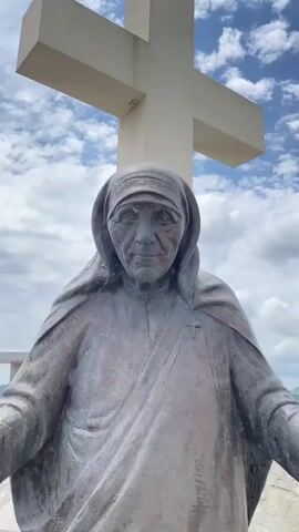 A video captures a bronze statue of an elderly woman, recognizable by her wimple as Mother Teresa. The statue features a serene, contemplative expression. In the background, a large, stark white cross stands against a bright blue sky dotted with fluffy white clouds. The camera slowly zooms in on the statue's face, highlighting the intricate details of the bronze work, including the wrinkles on her forehead and around her eyes. The setting appears to be outdoors, likely at a memorial or religious site, under clear daytime conditions. The location is Benkalj, Montenegro. No text is visible in the video.
