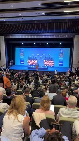 This media file captures a scene inside a large auditorium or conference hall, likely during a formal event. The setting is a stage area with a prominent blue backdrop displaying the "NATO OTAN ANKARA" logo, along with several American flags arranged in clusters. A podium with a seal, possibly presidential, stands center stage.

The foreground and midground are filled with rows of seated attendees, many of whom appear to be media personnel, evident by the numerous professional cameras and tripods set up throughout the audience. The individuals in the audience are diverse in age and gender, and their postures suggest they are observing the proceedings on stage, with some holding phones and cameras.

The activity taking place is likely a press conference, speech, or formal address related to NATO. The atmosphere appears professional and focused, with no overt displays of strong emotion. The lighting suggests it is daytime, possibly late morning or afternoon, as is common for such events. The mention of "Ankara" in the backdrop clearly indicates the location is in Ankara, Turkey, as per the prompt. The presence of multiple national flags suggests an international gathering.