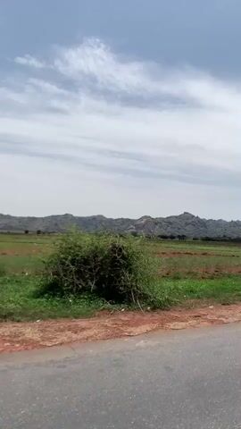 The video shows a landscape with a road in the foreground and a field leading up to a distant rocky hill range under a cloudy sky. The road is asphalt, gray in color, and has white lane markings. Adjacent to the road is a patch of red soil. The field is a mix of green grass and dry, brown patches. In the background, a series of hills or mountains are visible, characterized by large, gray rock formations. The sky overhead is partly cloudy, with a mix of blue sky and white clouds. The camera appears to be moving slowly along the road, and a side mirror is briefly visible in the lower right corner of the frame.