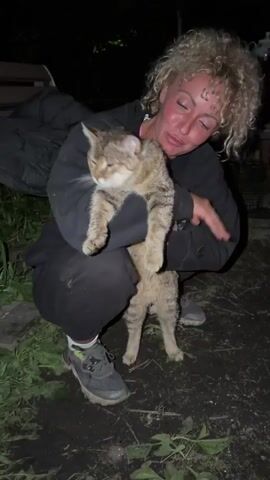 A person is crouched down, holding a light brown tabby cat. The person is fair-skinned, with curly blonde hair, and wearing a dark-colored hooded sweatshirt, dark-colored pants, and dark-colored athletic shoes with green accents. The person is looking at the cat and speaking to it, and seems to be affectionate towards it, even kissing the cat on the head. The cat is standing on its hind legs, held in the person's arms. The background is dark, but there is some grass and debris on the ground, and a wooden bench is visible in the background.