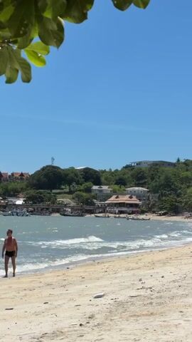 This media file captures a serene beach scene in Baan Bang Rak, Thailand. The foreground is dominated by a sandy beach, showing signs of gentle waves lapping at the shore. In the mid-ground, a calm bay is dotted with several small boats, suggesting a fishing or leisure community. A line of buildings, some with traditional roofs and others more modern, runs along the coast, interspersed with lush green trees. Beyond the buildings, a densely wooded hillside rises, topped with houses and a prominent telecommunications tower, indicating a settled but somewhat elevated residential area.

The sky is a clear, bright blue, signaling a sunny day, likely midday or early afternoon given the strong light and shadows. The overall mood is peaceful and idyllic, typical of a tropical coastal setting. There is no visible text in the media file. The scene suggests a tranquil day, possibly a holiday or a quiet moment for local residents.