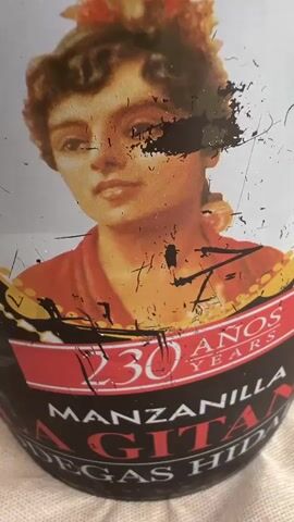 The video shows a close-up of a decorative bucket with a portrait of a woman and text that reads "230 AÑOS YEARS MANZANILLA SAN GINES BODEGAS HIDALGO". The bucket appears to be a wine or champagne cooler filled with ice. A clear glass bottle, containing a pale yellow liquid and labeled with red lettering, is partially submerged in the ice. A stemmed glass, also containing liquid, rests on top of the bottle. The background is blurred but suggests an indoor setting with a white tablecloth. The bucket has some wear and tear, with scratches and peeling paint.