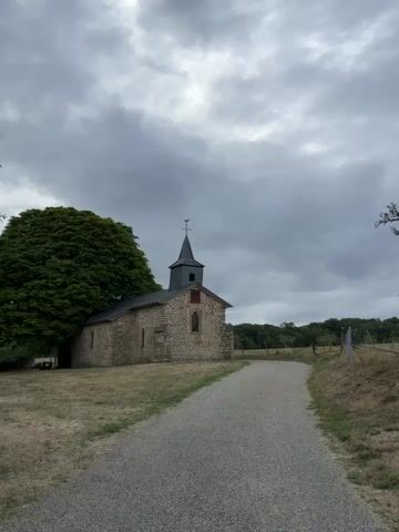 A gravel path leads towards a small, stone chapel under a cloudy sky. The chapel has a dark, pointed steeple with a weather vane and a single arched window on its facade. To the left of the chapel, a large, leafy tree provides shade. The surrounding area is a dry, grassy field, with a wooden fence visible in the distance. The camera pans slightly to the left, revealing more of the tree and the chapel's side.