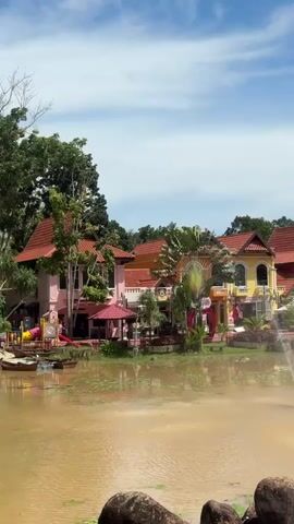 This video shows a tranquil scene with a muddy pond in the foreground, dotted with lily pads and edged by rocks. A fountain sprays water in the center of the pond. In the background, there are several colorful buildings with red tiled roofs, surrounded by lush green trees. The sky above is blue with scattered white clouds. The overall impression is of a peaceful, picturesque landscape.