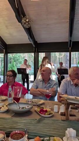 This is a vertical video of a band performing for a group of people seated at tables in a restaurant or event space. A female vocalist in a white suit is singing into a microphone, backed by several male musicians. One musician plays a saxophone, another a trumpet, and two others appear to be playing guitars. In the foreground, three men and one woman are seated at a table. The food on the table includes a salad, pickled vegetables, and bread. The background shows trees visible through large windows. The lighting is dim, with decorative light fixtures visible on the ceiling. The atmosphere appears to be a celebratory or formal gathering.