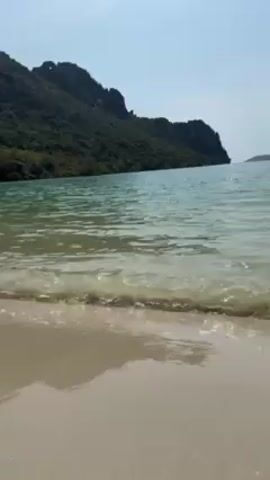 The video shows a scenic view of a beach. The sand is light in color, and the water is clear and turquoise. Small waves gently wash up on the shore. In the background, a green, mountainous island is visible against a clear, light blue sky. The scene is peaceful and serene.