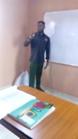 A man stands in front of a whiteboard in a room. He is wearing a dark long-sleeve shirt, green pants, and dark shoes. He holds a small dark object in his left hand and gestures with his right hand as he speaks. Behind him is a whiteboard with drawings on it. In front of him is a wooden desk with open books and papers on it. The walls of the room are light peach.