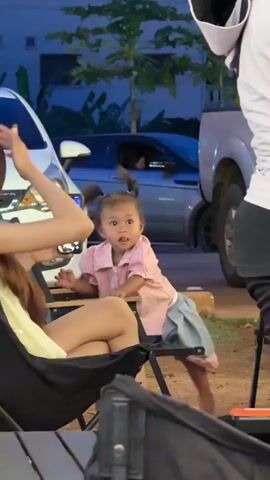 A young child with dark hair in pigtails, wearing a pink collared shirt and grey shorts, stands next to a chair. The child looks towards the camera with a surprised expression. A person's legs, wearing a white shirt and black pants, are visible on the right side of the frame. In the background, cars are parked and trees are visible. The setting appears to be outdoors, possibly at dusk.