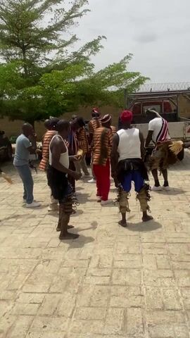 A group of about a dozen African men are dancing in a circle on a brick-paved area outdoors. Some of the men are wearing white tank tops and colorful shorts, often adorned with furry or fringed leg coverings and small jingle bells on their ankles. Others are in striped or patterned shirts and trousers. One man has a red cap. Most of them have their backs to the camera. They appear to be performing a traditional or celebratory dance, judging by their attire and movements. Several men on the edge of the group are wearing casual clothes. A truck, some trees, and a barbed wire fence are in the background. The sky is overcast.