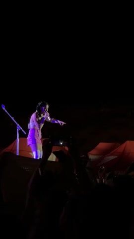 A nighttime outdoor concert is depicted from the perspective of a person in the crowd. Red tents with white logos are visible in the background. Many people are holding up their phones, their screens illuminated, and some are waving their arms. The crowd is silhouetted against the night sky and the stage lights. Some distant lights are visible in the upper portion of the frame.