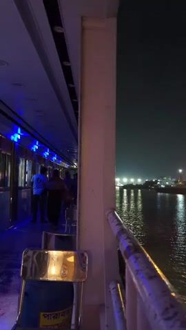 A nighttime view from the deck of a moving boat. The camera is angled slightly downwards towards a metallic railing. To the left, a walkway is illuminated by blue strip lights, with several people walking along it. Further down the walkway, more lights are visible. In the distance, across a body of water, a city skyline is visible with numerous bright lights. The water is dark and reflective, with small waves causing ripples. A white pillar is in the foreground, obscuring part of the walkway. A metallic chair with a blue seat is in the lower foreground. The sky is dark and clear.