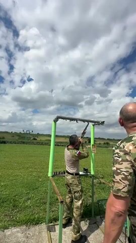 The video shows two men at an outdoor shooting range. One man, dressed in camouflage attire, is actively engaged in shooting. He is positioned behind a shooting stand equipped with a metal frame and green padding, aiming a shotgun upwards. His stance and focus suggest he is participating in a clay pigeon shooting event. The second man, also in camouflage, stands to the right, partially in view, seemingly observing or instructing.

The scene is set in a rural, grassy field under a dynamic sky filled with large, fluffy white clouds against a bright blue backdrop. The presence of a shooting apparatus, targets (though not clearly visible in action), and the activity itself point to a sporting or recreational shooting event. The overall atmosphere appears to be one of focused activity within a natural, open environment. The time of day seems to be daytime, possibly late morning or afternoon, given the bright lighting. The weather appears to be fair, with the clouds suggesting a possibility of scattered showers but not overcast conditions. The location, indicated by "Фельдмаршальский, Russia," places the event in a specific rural area in Russia. No discernible text is visible in the video.